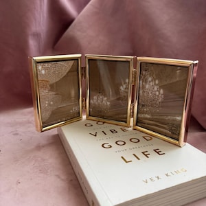 May include: A gold-coloured, three-panel picture frame is open, displaying sepia-toned images. The frame sits atop a white book with gold lettering that reads "GO VIBE GOOD LIFE". The background is a pink fabric.