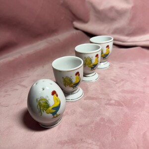 May include: A set of four white ceramic egg cups and a matching salt shaker. Each piece is decorated with a colourful rooster design. The egg cups have a gold rim and a pedestal base. The salt shaker is egg-shaped with holes on top.