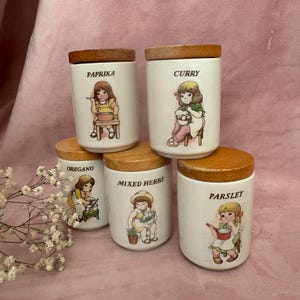 May include: A collection of six white ceramic spice jars with wooden lids. Each jar is labelled with a spice name and a cartoon illustration of a child. Spices include paprika, curry, oregano, mixed herbs, and parsley.