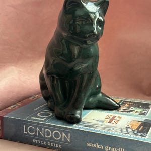 May include: A dark green ceramic cat figurine in a seated position. The cat is glossy and has a smooth finish. It sits atop a book titled "LONDON STYLE GUIDE" with a Union Jack stamp design.