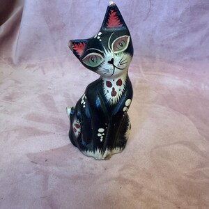 May include: A black and white cat figurine with red and white accents. The cat has green eyes and a whimsical design. The figurine is hand-painted and has a vintage aesthetic. The cat is sitting upright with its head tilted.