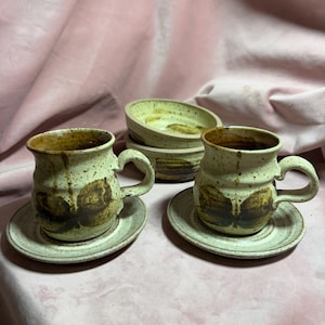 May include: A handmade ceramic coffee cup and saucer set. The cups are speckled beige with brown accents and a handle. The saucers are a matching speckled beige. A stack of two small bowls with a similar design is also visible.