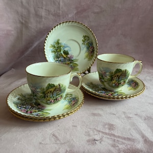 May include: A set of vintage teacups and saucers. The cups and saucers are cream-colored with a gold trim and a landscape scene. The set includes two cups, two saucers, and one additional saucer.