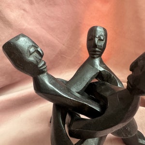 Puede incluir: Una escultura de madera marrón oscuro que representa tres figuras humanas estilizadas entrelazadas. Las figuras tienen cuerpos simples y alargados y rasgos faciales tallados. La escultura está sobre un fondo rosa.