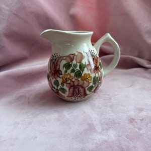 Op de afbeelding: Crèmekleurige keramische creamer met een bloemenpatroon in bruin, geel en groen. Het heeft een handvat en een tuit. Het bloemmotief loopt rond de body van de creamer. Een decoratief item.
