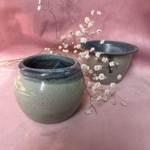 Può includere: Due vasi in ceramica con smalto verde chiaro e blu. Il vaso rotondo ha un'ampia apertura e un bordo blu scuro. L'altro vaso ha una forma più piccola e affusolata con un bordo blu simile. Fiori bianchi disposti tra i vasi su sfondo rosa.
