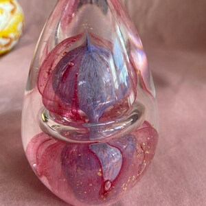 May include: A clear glass paperweight in a teardrop shape. It features a swirling design of pink and purple glass with gold flecks. A clear glass ring encircles the centre. The paperweight is displayed on a pink surface.