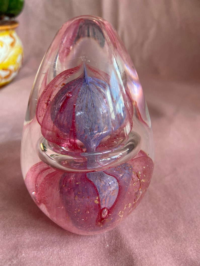 May include: A clear glass paperweight in a teardrop shape. It features a swirling design of pink and purple glass with gold flecks. A clear glass ring encircles the centre. The paperweight is displayed on a pink surface.