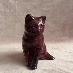 May include: A ceramic cat figurine in a sitting position. The cat is a deep burgundy color with black marbling throughout. The figurine has a glossy finish and is set against a pink background.