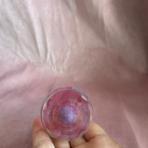 May include: A clear glass object with a swirling interior of red, purple, and gold colours. The glass has a rounded shape and is held in a hand, with a pink background.