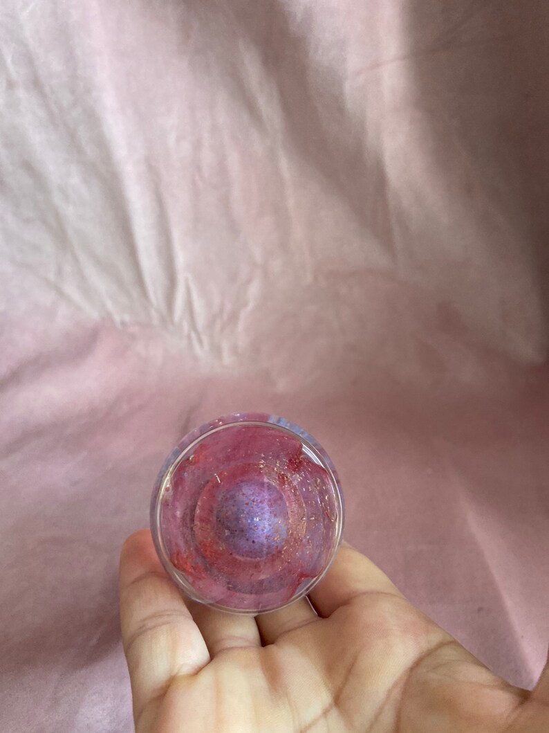 May include: A clear glass object with a swirling interior of red, purple, and gold colours. The glass has a rounded shape and is held in a hand, with a pink background.