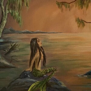 May include: A mermaid with long dark hair sits on a rock with her tail in the water. The water is a light blue and the sky is a soft orange. The background is a blurry image of trees and a lake.