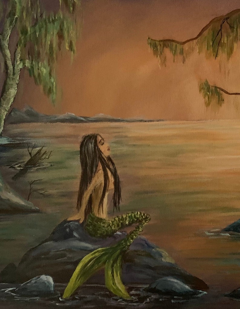 May include: A mermaid with long dark hair sits on a rock with her tail in the water. The water is a light blue and the sky is a soft orange. The background is a blurry image of trees and a lake.