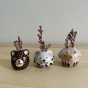 Puede incluir: Tres pequeños maceteros de cerámica con forma de animal con flores secas. Los maceteros tienen forma de oso, gato y perro. El macetero de oso es marrón con lunares dorados, el macetero de gato es blanco con manchas negras y orejas rosas, y el macetero de perro es rosa con detalles verdes y dorados.