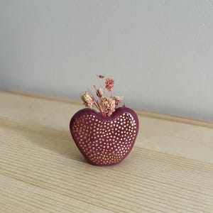 Peut inclure: Un petit vase en forme de cœur avec une finition bordeaux et des pois dorés. Le vase est rempli de fleurs roses séchées.