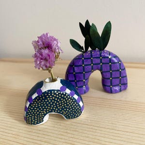 Puede incluir: Dos pequeños jarrones de cerámica con forma de arco, uno con un patrón de cuadros morados y dorados y el otro con fondo blanco, detalles en azul marino y lunares dorados. El jarrón con lunares tiene un pequeño ramo de flores moradas.