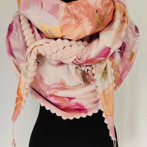 May include: A floral print scarf in shades of pink, peach, and gold, displayed on a black mannequin. The scarf has a soft, textured trim and matching tassels. The design features a watercolor-style floral pattern.