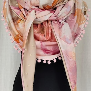 May include: A floral print scarf with pink, peach, and beige tones, adorned with pink pom-poms and tassels. The scarf is draped over a black mannequin, showcasing its design and texture.