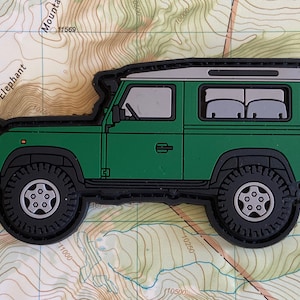 以下が含まれることがあります： 黒の輪郭と白い屋根を持つ緑のランドローバーディフェンダーのパッチ。パッチは銀色のリムが付いた黒いタイヤを持っています。