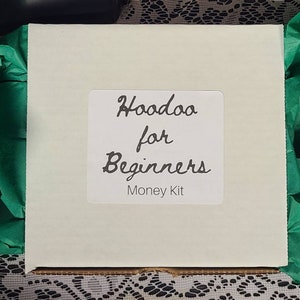 Può includere: Una scatola quadrata bianco sporco, etichettata "Hoodoo for Beginners Money Kit", è appoggiata su carta velina verde. La scatola è circondata da altra carta velina verde e poggia su una superficie di pizzo bianco.