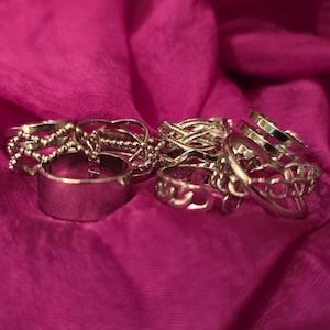 May include: A set of silver rings with different designs, including a thick band, a braided ring, and a ring with a heart-shaped design. The rings are arranged on a pink fabric background.