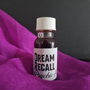 Pode incluir: Uma pequena garrafa de vidro com uma tampa branca e um rótulo que diz "DREAM RECALL Psychic S". A garrafa está cheia de um líquido roxo escuro e está sobre um tecido roxo.