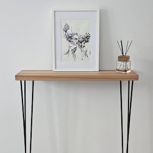 Op de afbeelding: Een houten console met een lichtbruin blad en zwarte haarspeldpoten. Een ingelijste botanische print en een glazen diffuser met riet staan op de tafel. De tafel is ongeveer 76 cm lang.