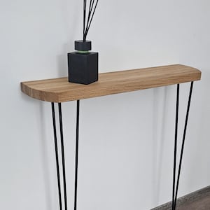 Peut inclure: Une console moderne et minimaliste avec un plateau en bois naturel et des pieds en épingle noirs. Un flacon diffuseur noir avec des roseaux est posé sur la table. La table mesure environ 100 cm de long.