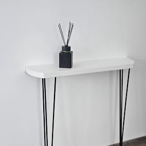 Puede incluir: Una consola blanca con patas negras de horquilla. Un difusor cuadrado negro con varillas se encuentra sobre la mesa. La mesa está contra una pared blanca y un suelo de imitación madera.