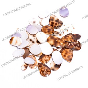 May include: A collection of polished gemstones in various shapes and colors. The gemstones include white, brown, and lavender hues, with some featuring speckled patterns. The assortment is scattered on a white background, showcasing the natural variations in the stones.