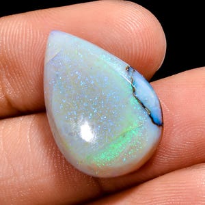 Può includere: Una gemma di opale a forma di goccia, prevalentemente bianca con riflessi iridescenti blu e verdi. Una vena marrone scuro attraversa la pietra. L'opale è tenuto in una mano.