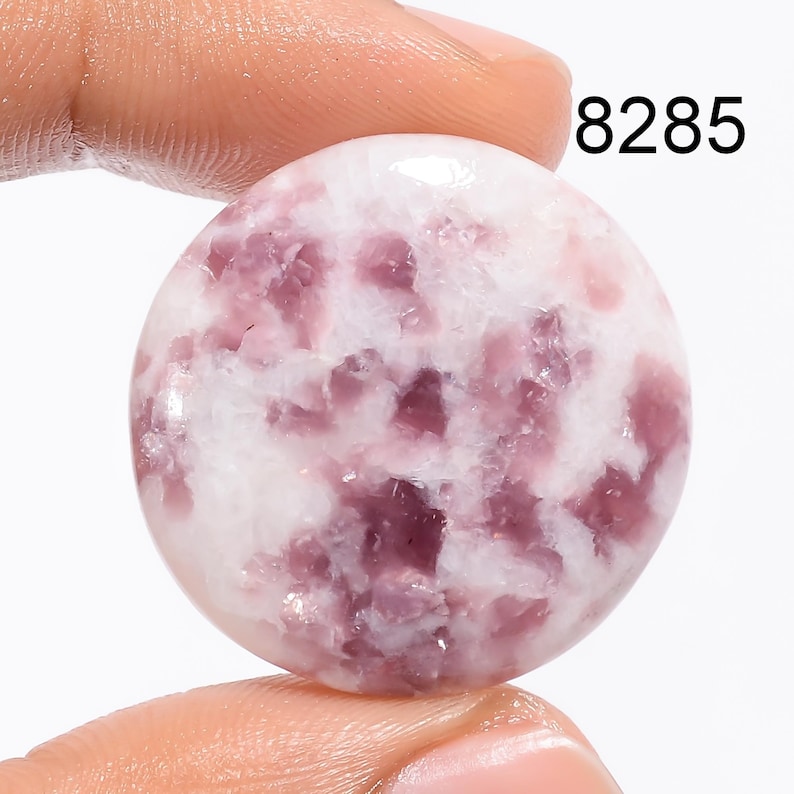 May include: A round, white cabochon gemstone with pink and purple inclusions. The stone is approximately 1 inch in diameter.