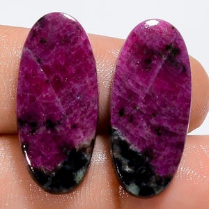 May include: Two oval-shaped cabochons featuring a vibrant magenta and deep pink color palette with distinct black inclusions at the base. These polished gemstones display a smooth, reflective surface with natural crystalline textures throughout.