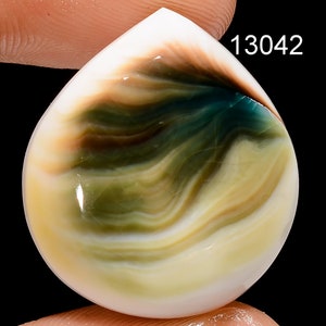 May include: A teardrop-shaped cabochon gemstone with swirling patterns of white, brown, green, and blue. The stone has a smooth, polished surface.