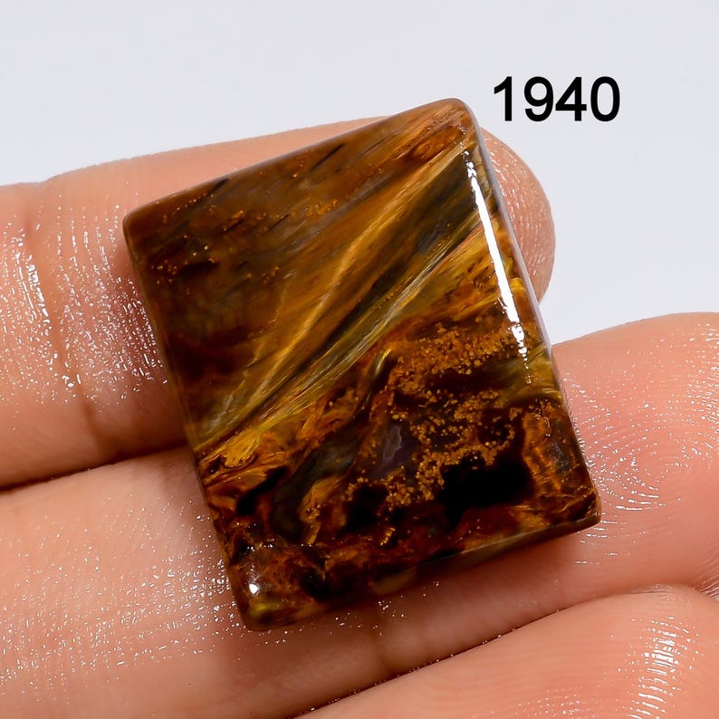 K&ouml;nnte beinhalten: Ein brauner, goldener und schwarzer Tigerauge-Cabochon mit rechteckiger Form. Der Edelstein hat eine polierte, glatte Oberfl&auml;che und ein einzigartiges, wirbelndes Muster.