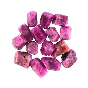 May include: A cluster of raw ruby gemstones, each with a rough, irregular shape. The rubies display a vibrant pinkish-red hue, with some stones showing darker inclusions. The gemstones are scattered against a plain white background.