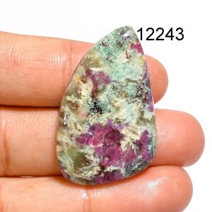 May include: A polished gemstone with a teardrop shape. The stone features a mix of colours, including deep magenta, light green, and cream. The number "12243" is printed in black on the upper right side of the stone. The stone is held in a hand.