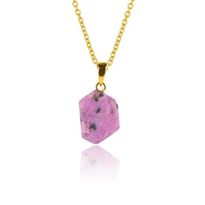 May include: A gold chain necklace featuring a pink, hexagonal pendant. The pendant is a rough-cut gemstone with dark flecks. The necklace is set against a white backdrop, with a reflection of the pendant below.