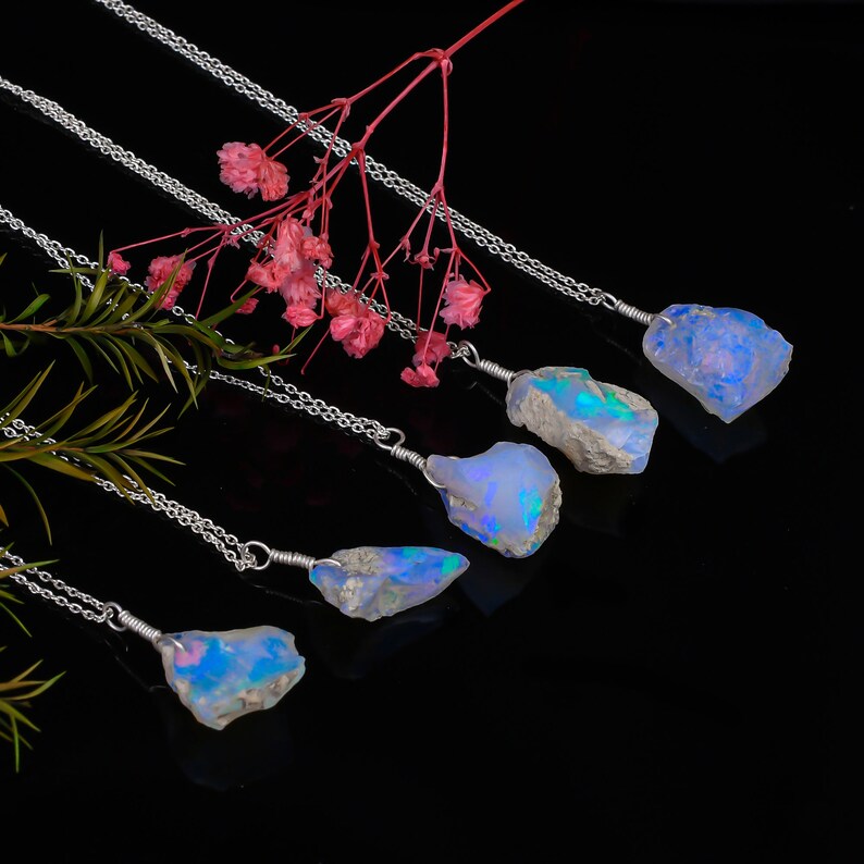 May include: Five silver necklaces with raw opal pendants. The opals are iridescent and have a blue, green, and purple hue. The necklaces are displayed against a black background.