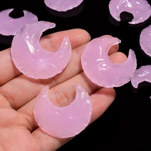 Puede incluir: Varias cuentas translúcidas en forma de media luna, de color rosa claro, se muestran sobre un fondo negro. Las cuentas tienen un aspecto liso y pulido y probablemente están hechas de vidrio o un material similar. Algunas se sostienen en una mano.