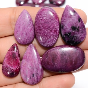 May include: A collection of five polished cabochon gemstones in shades of magenta, deep pink, and purple. The stones feature varied shapes including teardrops and ovals, with some displaying dark black inclusions and a smooth, reflective surface.