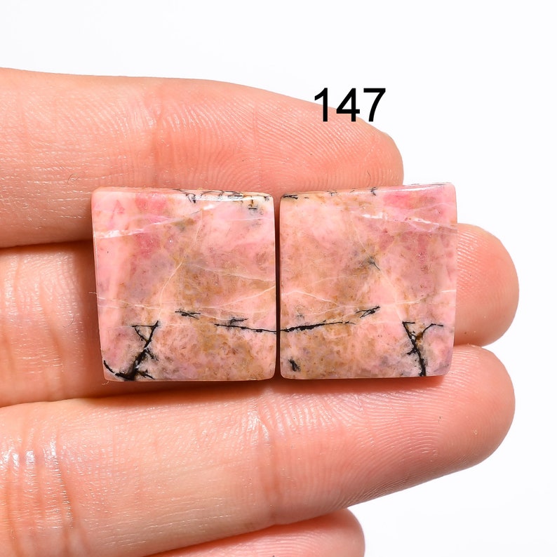 May include: Two square-shaped pink and brown rhodochrosite cabochons with black veins. The stones are approximately 1 inch in size. 147