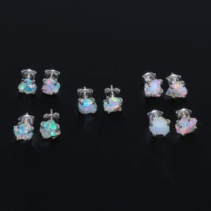 May include: Six pairs of opal stud earrings. Each earring features a raw, irregular-shaped opal stone in shades of white, blue, and pink. The opals are set on silver posts with butterfly backs. The earrings are displayed against a black background.