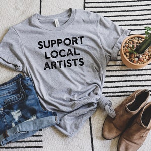 May include: A gray t-shirt with the text "SUPPORT LOCAL ARTISTS" printed in black. The shirt is laid out on a striped rug with a pair of brown ankle boots and a potted cactus.