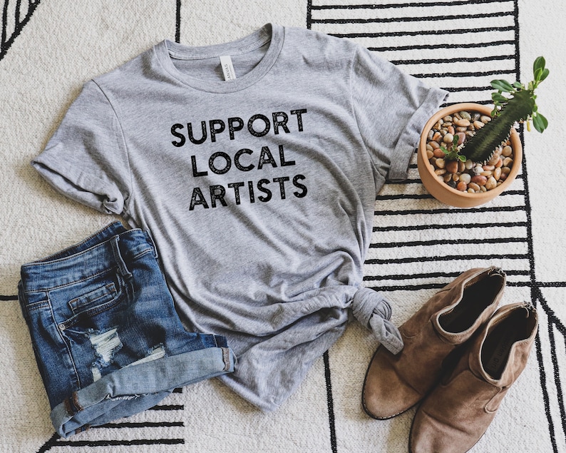 May include: A gray t-shirt with the text "SUPPORT LOCAL ARTISTS" printed in black. The shirt is laid out on a striped rug with a pair of brown ankle boots and a potted cactus.