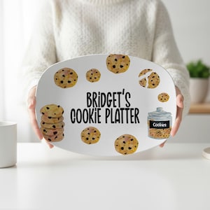 May include: An oval white serving platter featuring a watercolor-style illustration of chocolate chip cookies and a glass cookie jar. The text "BRIDGET'S COOKIE PLATTER" is printed in black in the center of the platter, surrounded by scattered cookies.