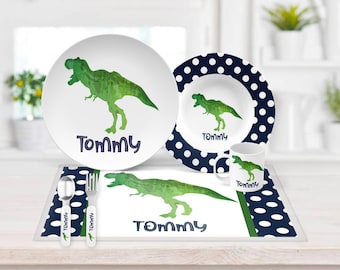 May include: A children's dinnerware set including a plate, bowl, mug, placemat, spoon, and fork. Each piece features a green watercolor-style T-Rex dinosaur and the name Tommy in dark blue text. The bowl and placemat have a navy blue polka dot border.