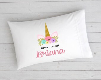 May include: A white rectangular pillowcase featuring a graphic of a unicorn face with a gold horn, pink ears, and a floral crown in shades of pink, blue, and purple. Below the unicorn graphic, the name Briana is printed in a pink cursive font.