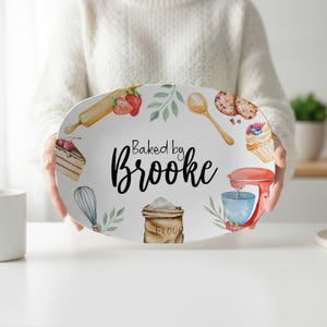 May include: An oval white serving platter featuring a watercolor illustration of baking tools and ingredients, including a rolling pin, whisk, flour sack, and stand mixer. The text "Baked by Brooke" is printed in black in the center.
