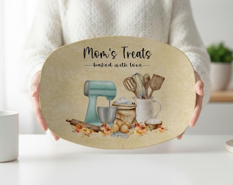 May include: An oval serving platter with a beige background featuring the text Mom's Treats baked with love. The design includes a light blue stand mixer, a bag of flour, a wooden spoon, a whisk, a rolling pin, eggs, and autumn-colored flowers.